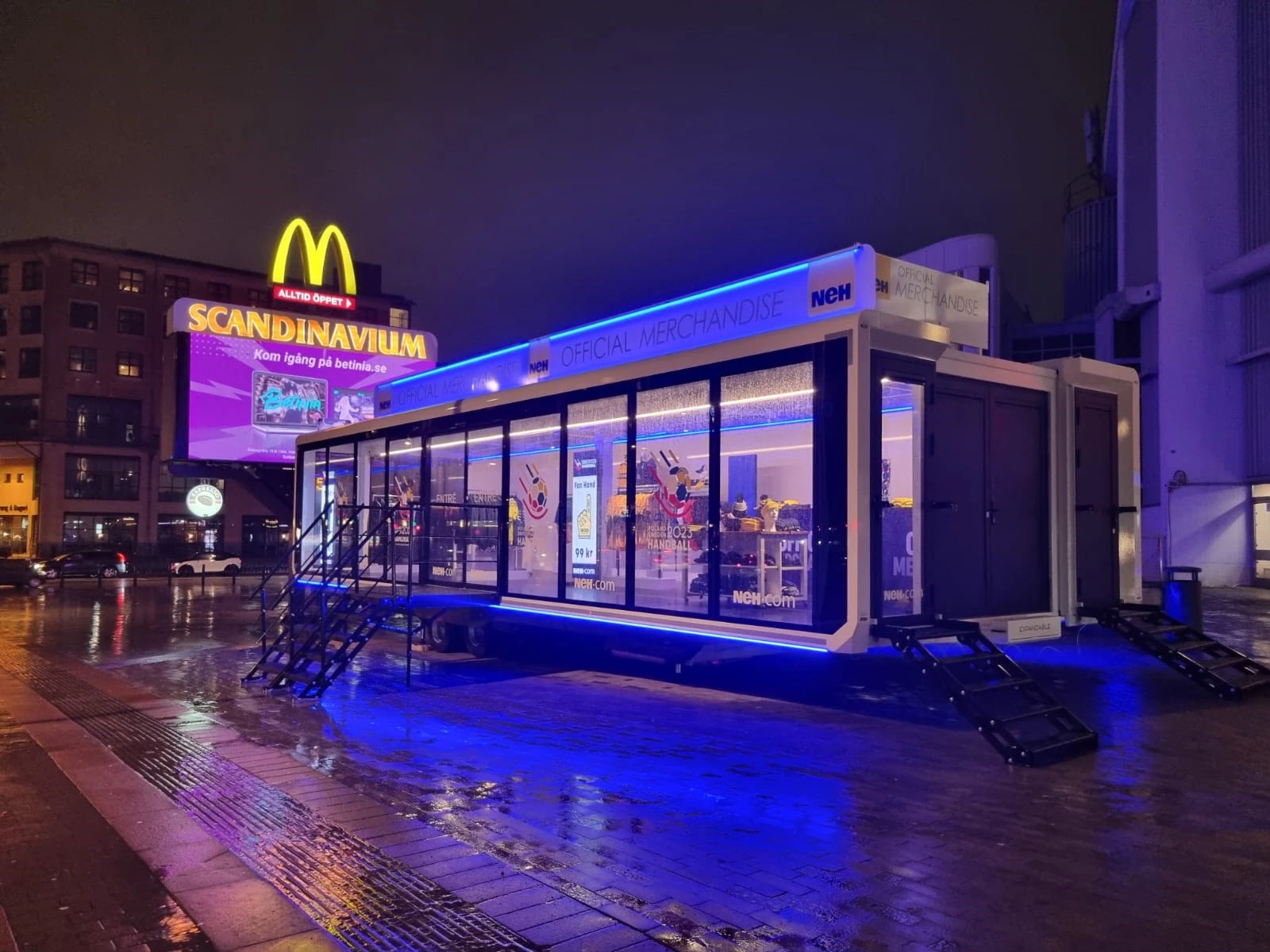 A blue-lit merchandise trailer on a rainy street at night, showcasing sports gear through glass walls.